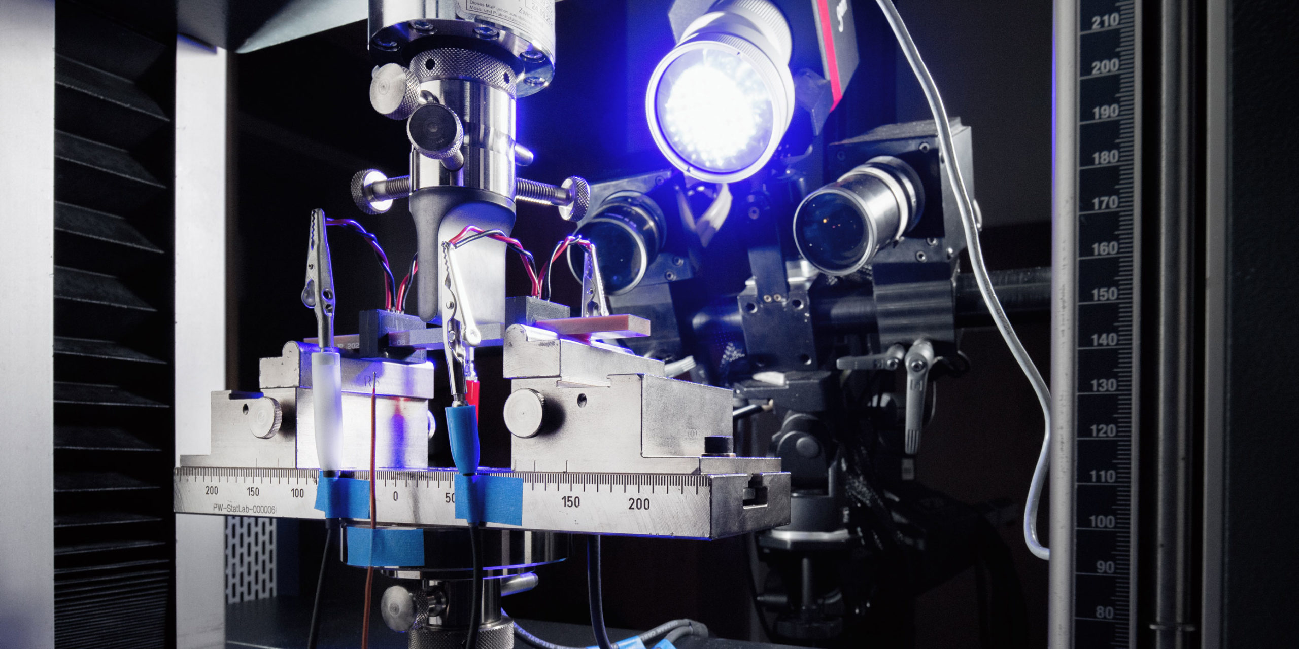 Glasfaserverstärkte Kunststoffprobe im Biegeversuch. Der Superkondensator ist mittig im Randbereich der Probe integriert. Die mechanische Messung wird durch eine elektrochemische Messung begleitet.