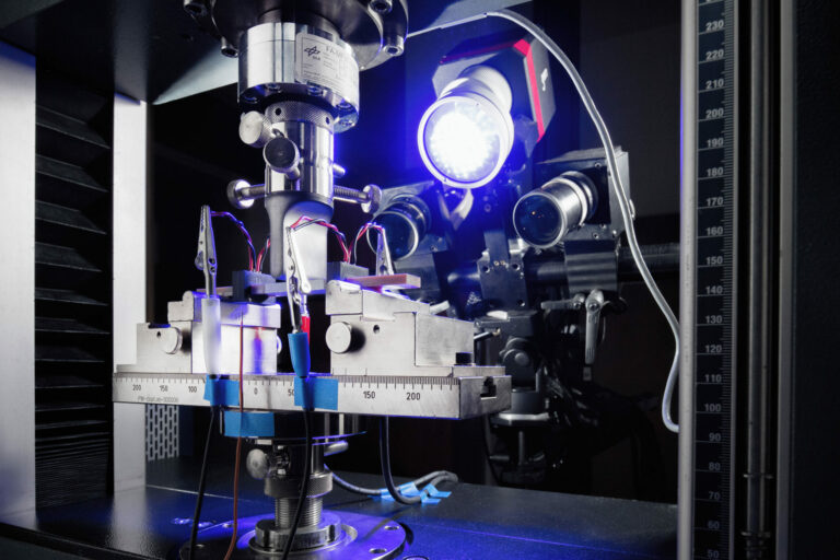 Glasfaserverstärkte Kunststoffprobe im Biegeversuch. Der Superkondensator ist mittig im Randbereich der Probe integriert. Die mechanische Messung wird durch eine elektrochemische Messung begleitet.